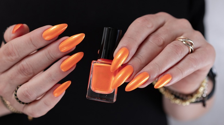 Manos con manicura naranja brillante sosteniendo una botella de esmalte de uñas naranja