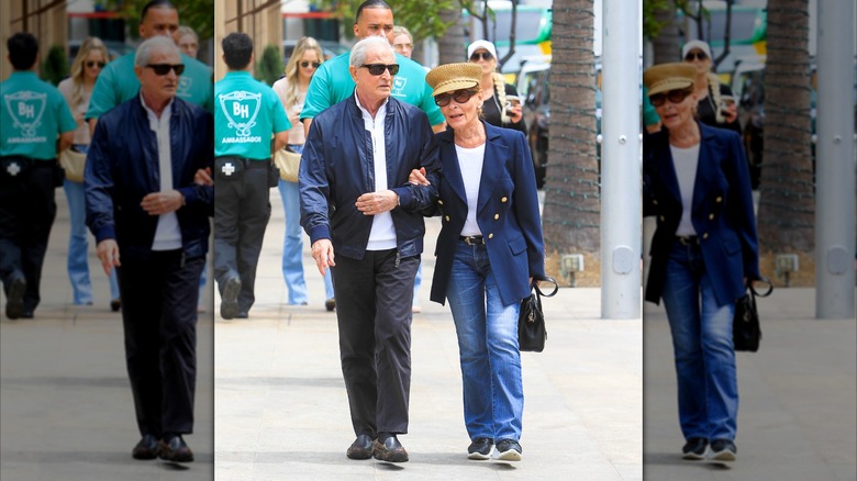 Judy Sheindlin camina con su esposo (izq.) vistiendo jeans acampanados y una manga larga blanca con una chaqueta azul y un sombrero de pescador.