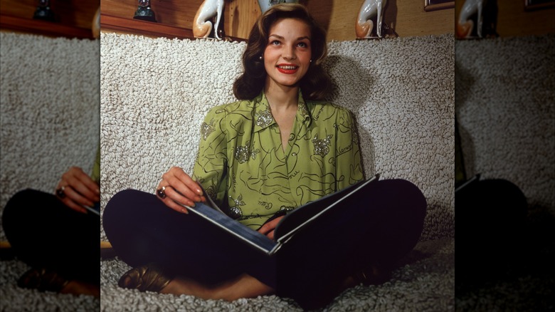 Lauren Bacall sentada con las piernas cruzadas con un libro abierto en su regazo, vestida con una blusa verde adornada y lápiz labial rojo, alrededor de 1945.