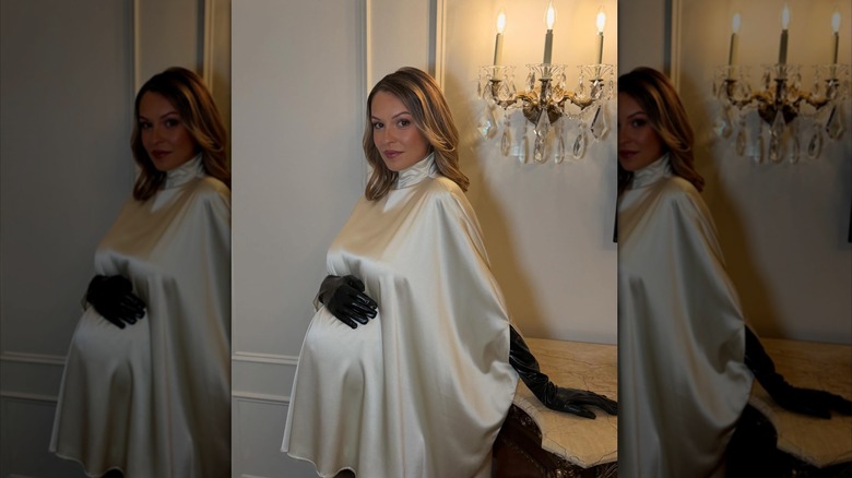 Kayla Wallace con un vestido de cuello alto de seda blanco y guantes de cuero negros con la mano en la panza.
