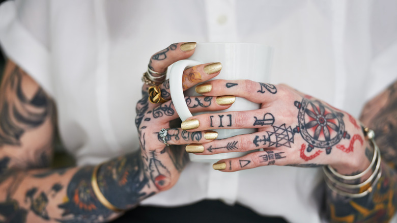 Una persona con manos y nudillos tatuados sosteniendo una taza.