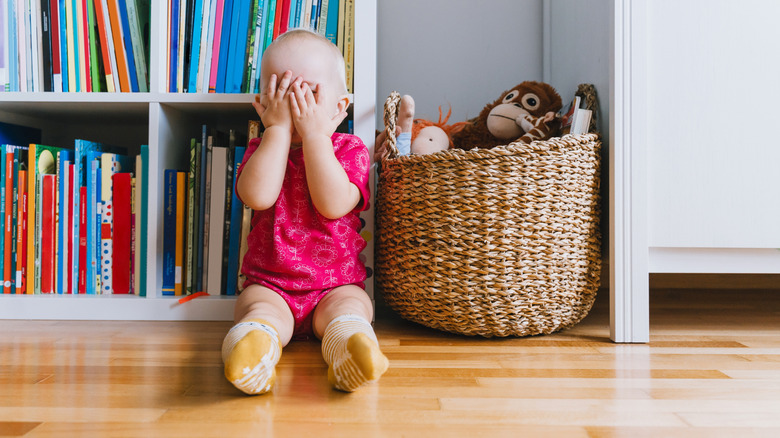 Una niña pequeña se sienta frente a una estantería colorida junto a una canasta de juguetes que le cubre la cara.